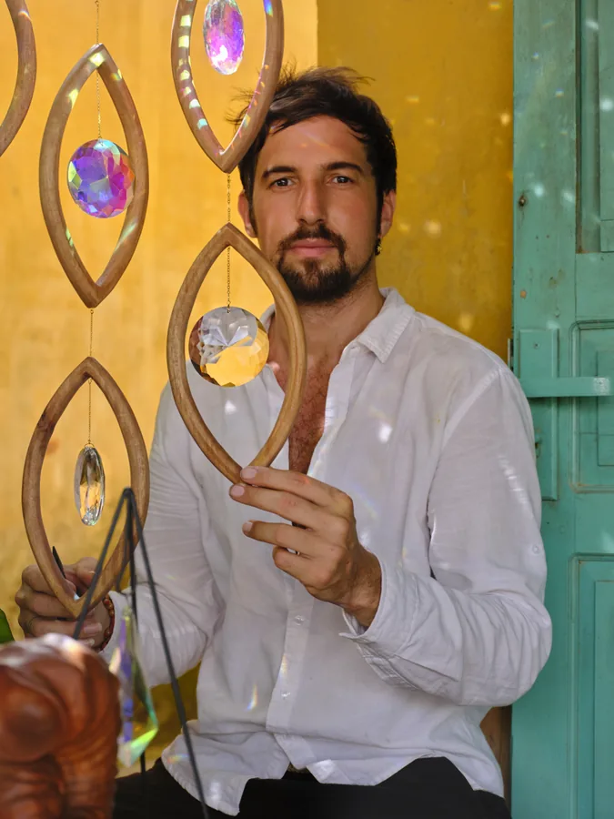 Portrait of Arihant Ortega in a white shirt, seated among hanging wooden eye pieces against a yellow wall