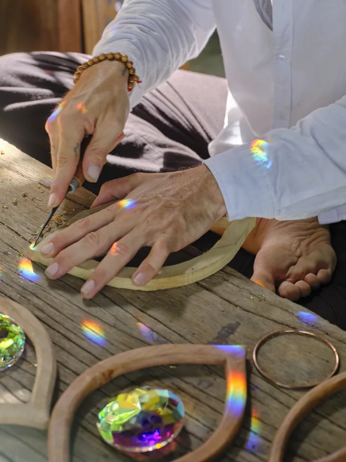 Hands shaping a carved wooden eye on a workbench, the spectrum from a crystal falling across the wood